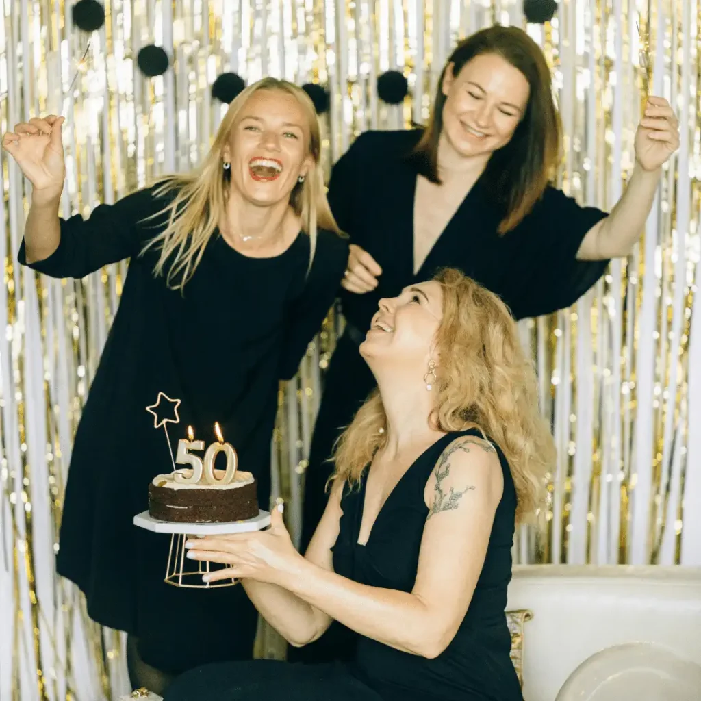 Three women in black dresses celebrating a 50th birthday with a lit cake and sparklers, highlighting joyful friendships and social connection in midlife healthy aging.