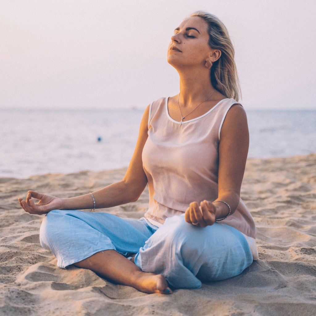 Woman in midlife meditating cross-legged on the beach at sunset, practicing mindful breathing for calm, healthy aging