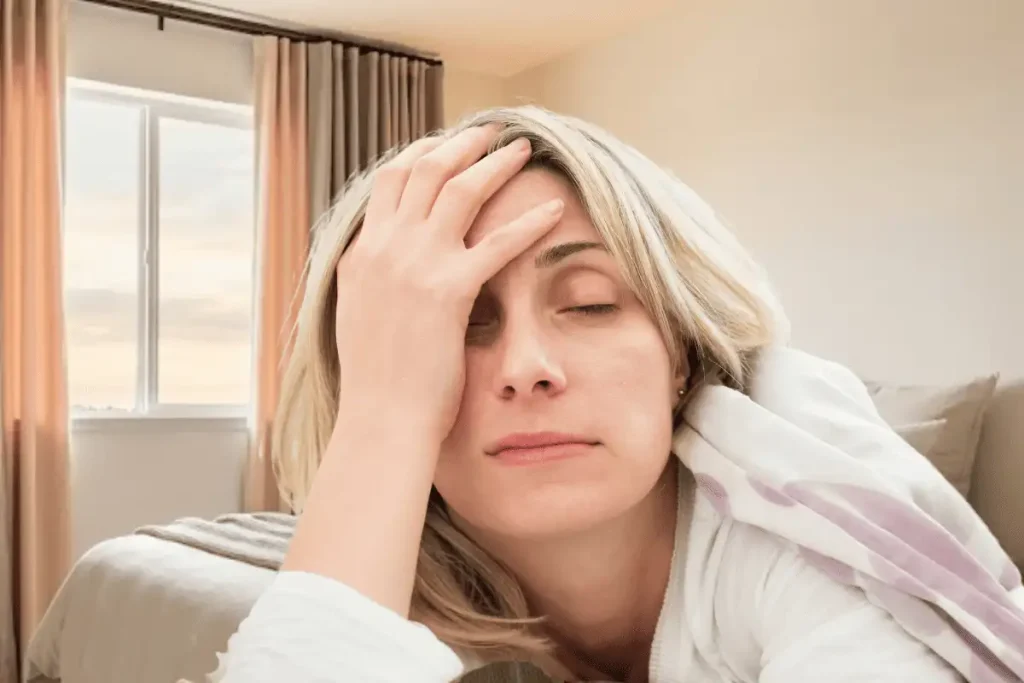 Tired midlife woman sitting on her bed with her hand on her head in a softly lit bedroom, illustrating trouble sleeping and fatigue after age 50.