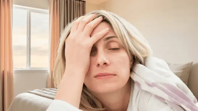 Tired midlife woman sitting on her bed with her hand on her head in a softly lit bedroom, illustrating trouble sleeping and fatigue after age 50.