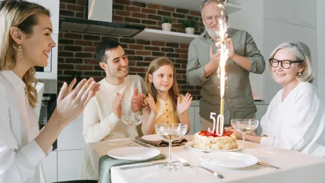 Family celebrating a woman’s 50th birthday around a dinner table with a cake and sparkling candle, symbolizing joyful, connected healthy aging.