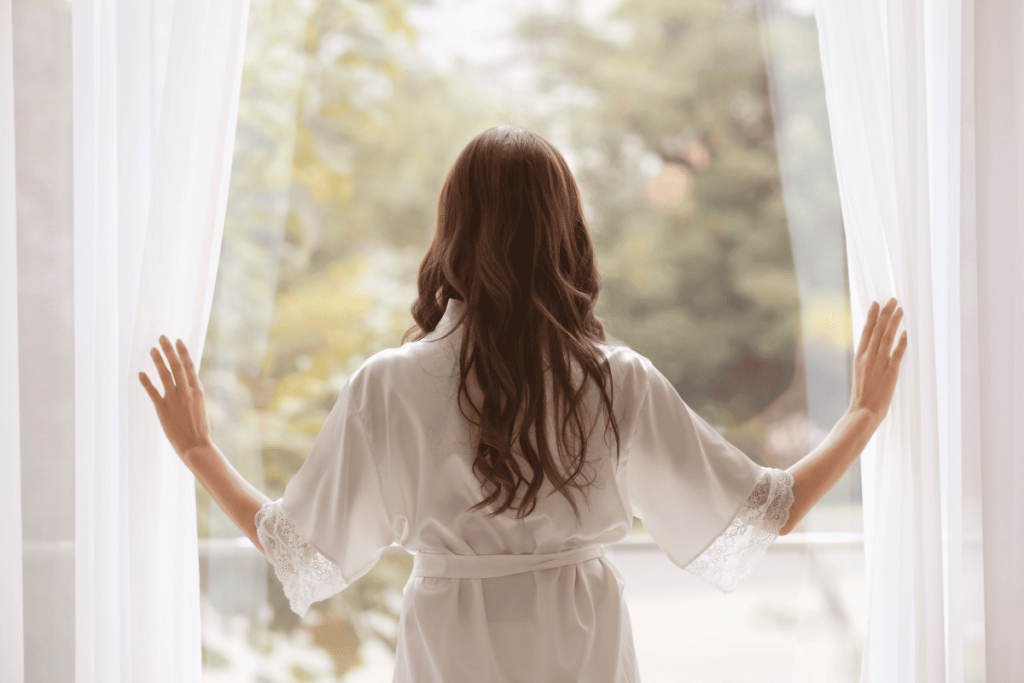 Woman in elegant white robe standing by window with soft morning light, representing healthy morning routine for menopausal vitamin D intake.