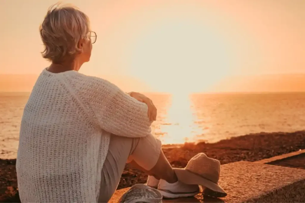 Woman over 50 sitting by the sea at sunrise, embracing a new life direction in a peaceful dawn setting.