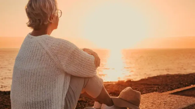 Woman over 50 sitting by the sea at sunrise, embracing a new life direction in a peaceful dawn setting.